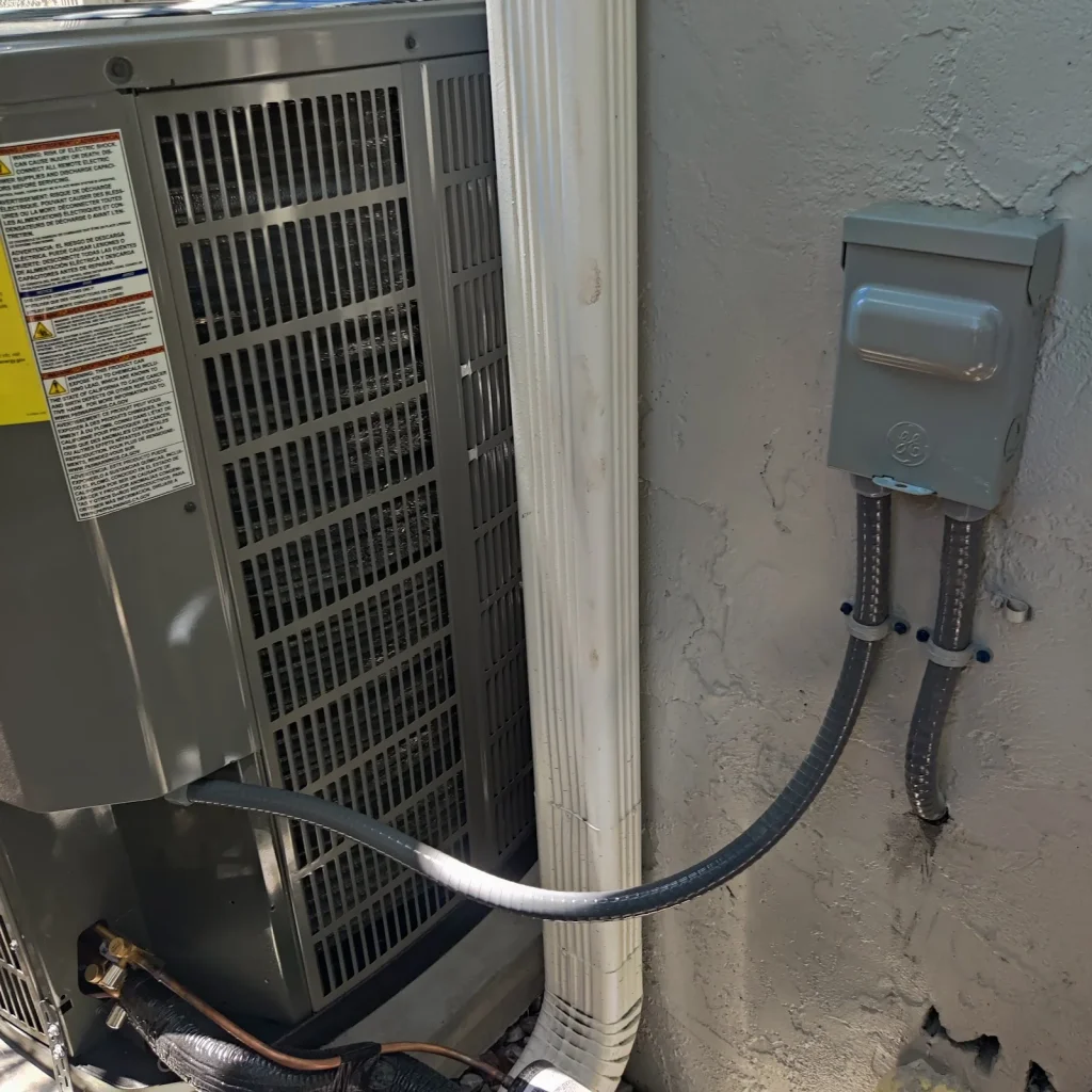 Gray outdoor AC disconnect box on a stucco wall with flexible conduit running to an air conditioner condenser at a Pinellas County home